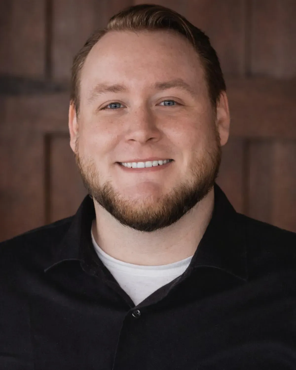 A person with a short beard and brown hair smiles while wearing a black collared shirt over a white undershirt.