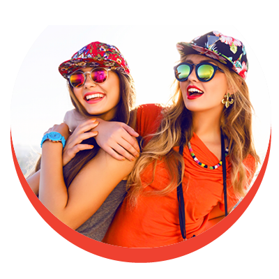 Two teenage girls in a playful embrace are sporting sunglasses, colorful hats and large smiles
