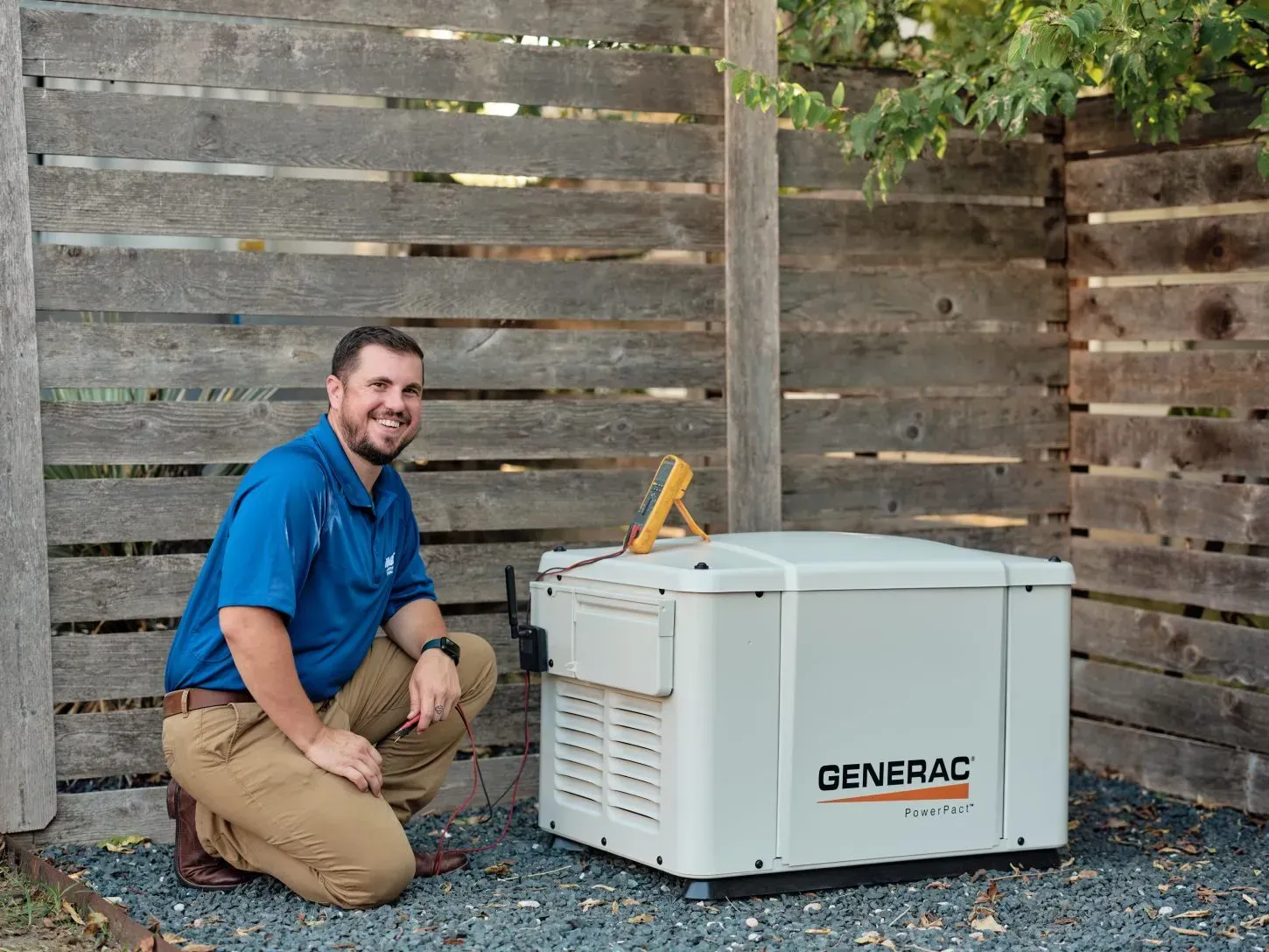 Man kneeling beside a white portable generator in a backyard, smiling near a wooden fence.