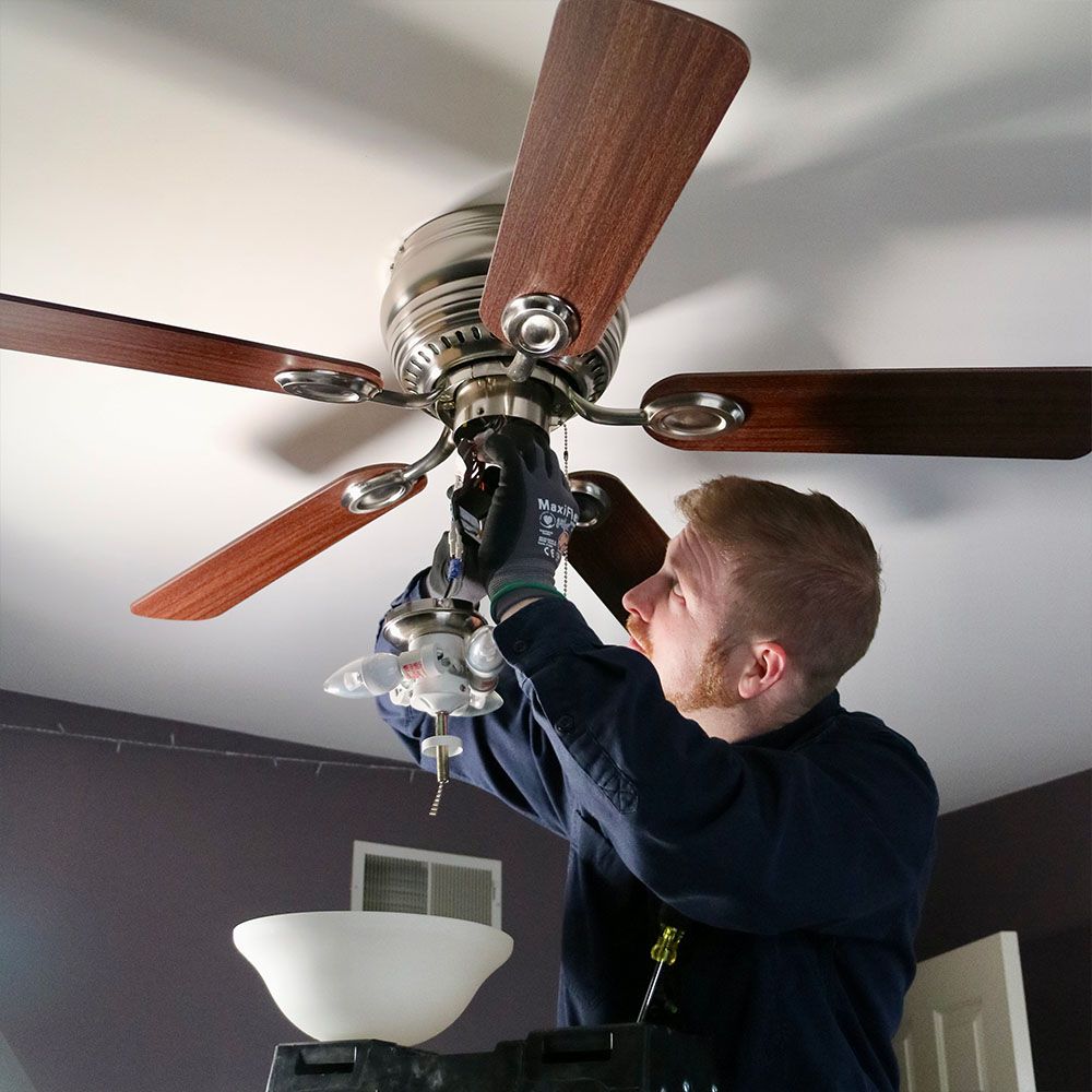 Man installing a ceiling fan with wooden blades in a room, reaching up with tools.