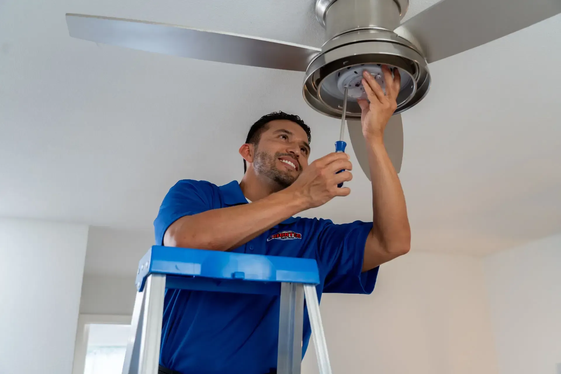 Electrician standing on a ladder repairing a ceiling fan with a screwdriver in a white room