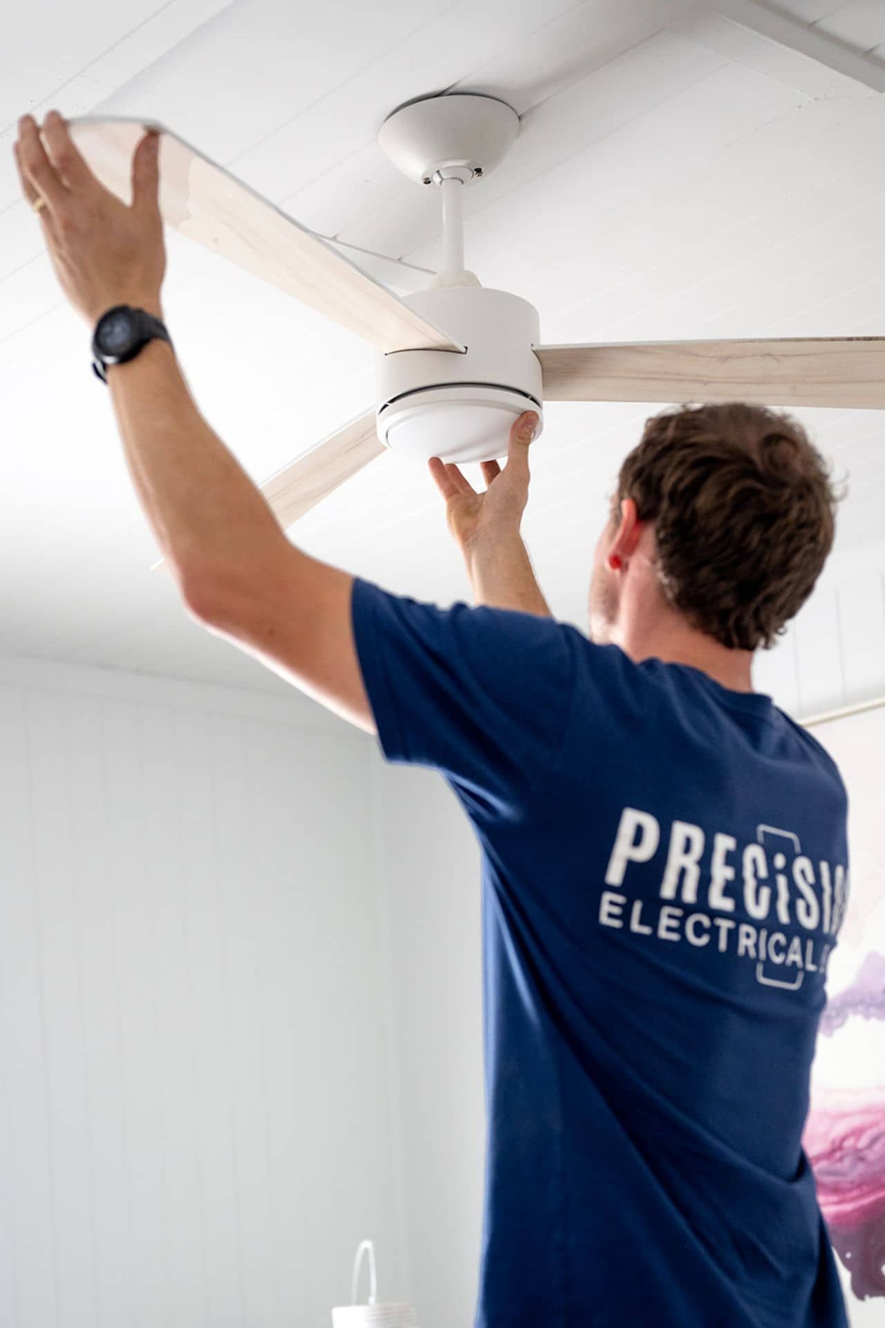 Electrician installing a white ceiling fan in a bright room, wearing a blue Precision Electrical shirt.