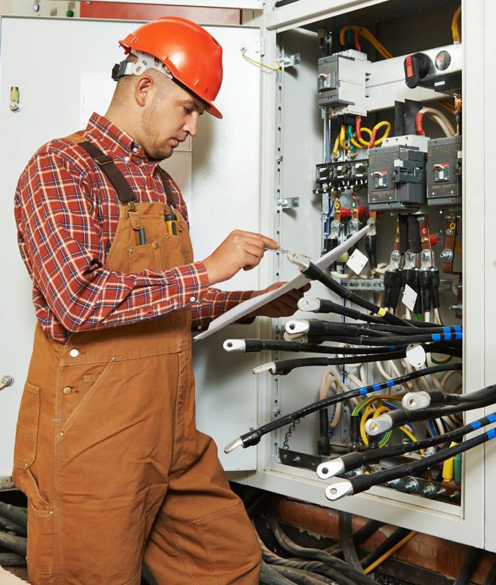 Electrician in orange hard hat inspecting an open electrical panel with tools in a utility room