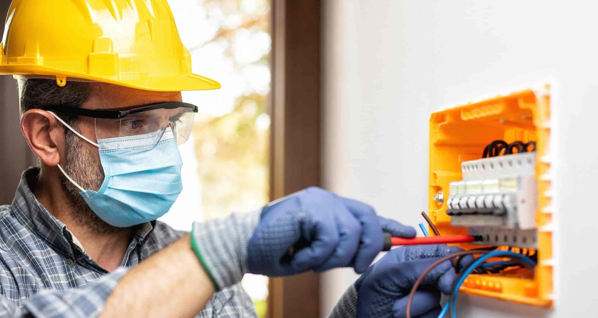 Electrician in hard hat and mask wiring an orange electrical panel with gloves