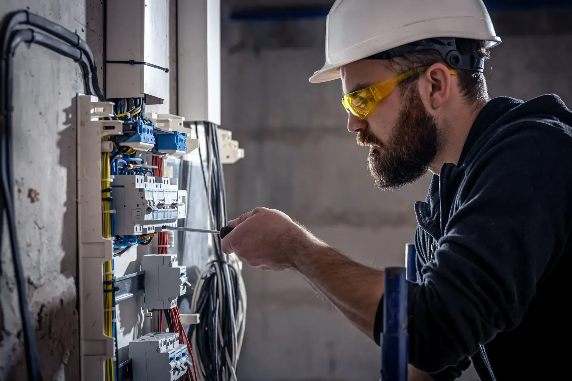 Electrician in hard hat and safety glasses wiring an electrical panel indoors