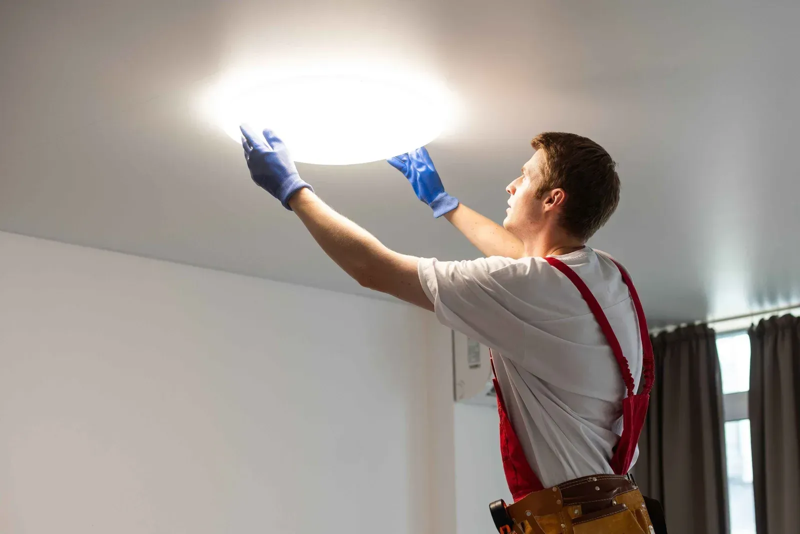 Technician in blue gloves reaching up to inspect or repair a ceiling light fixture indoors