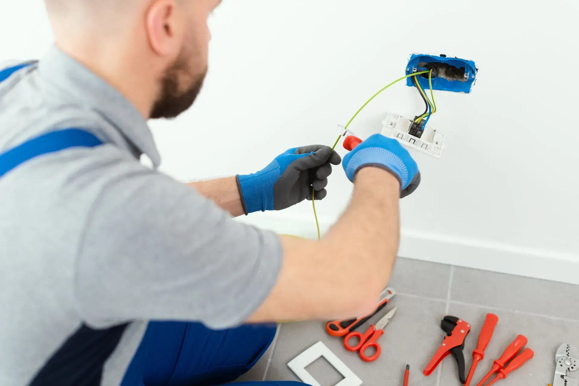 Electrician wiring a wall outlet with blue gloves and tools on the floor
