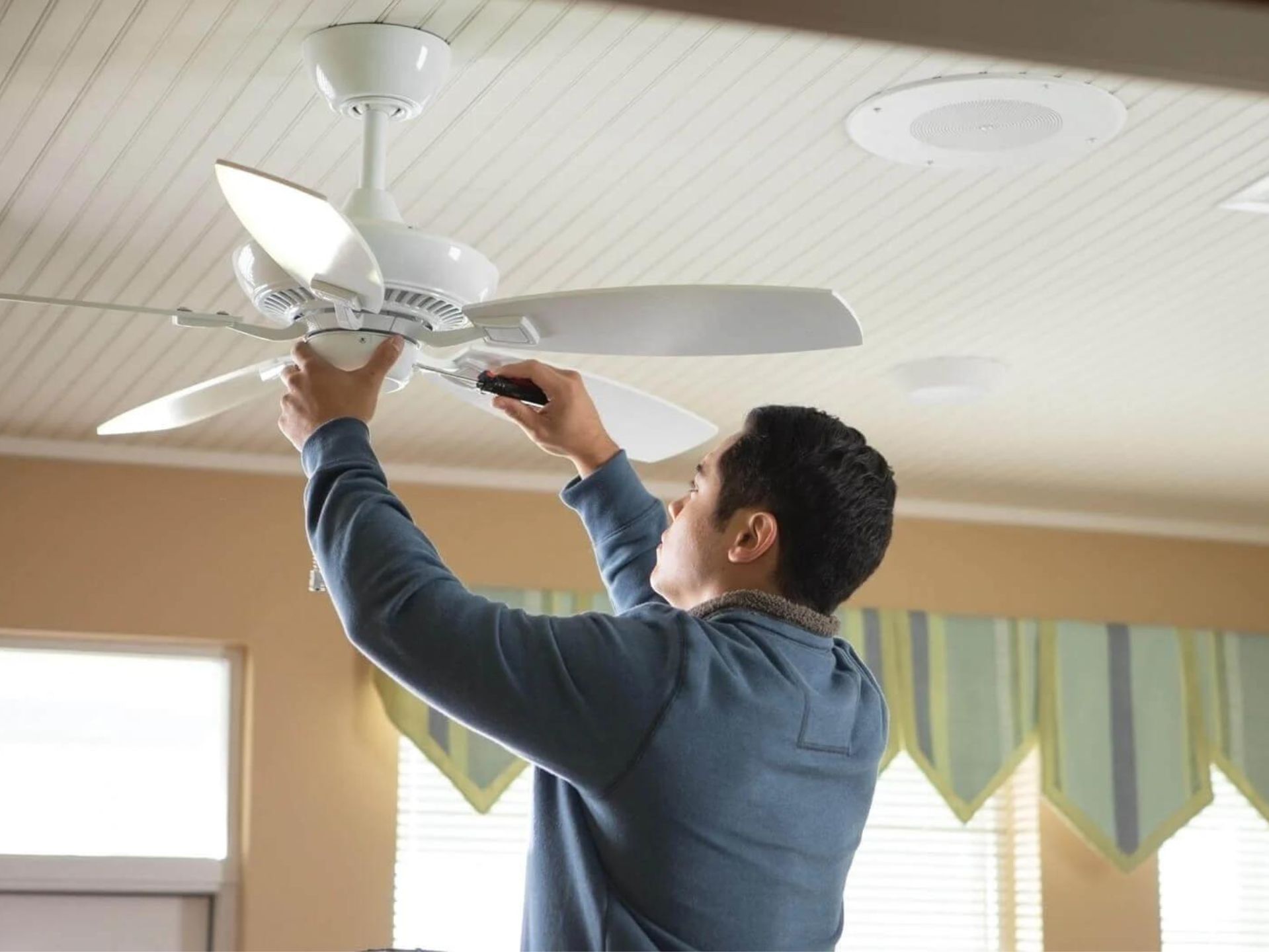 Man installing a white ceiling fan in a room with striped walls and a window