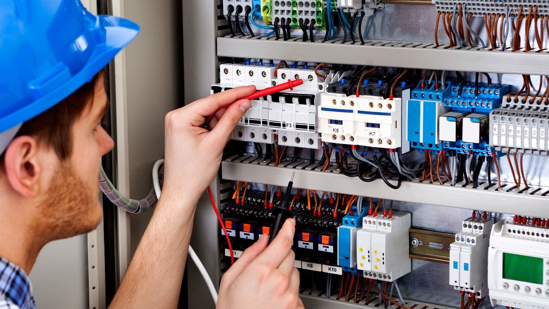 Technician in a blue hard hat testing an electrical control panel with a multimeter.