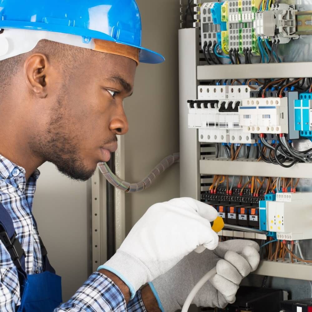 Technician in blue hard hat wiring an electrical control panel with tools and gloves