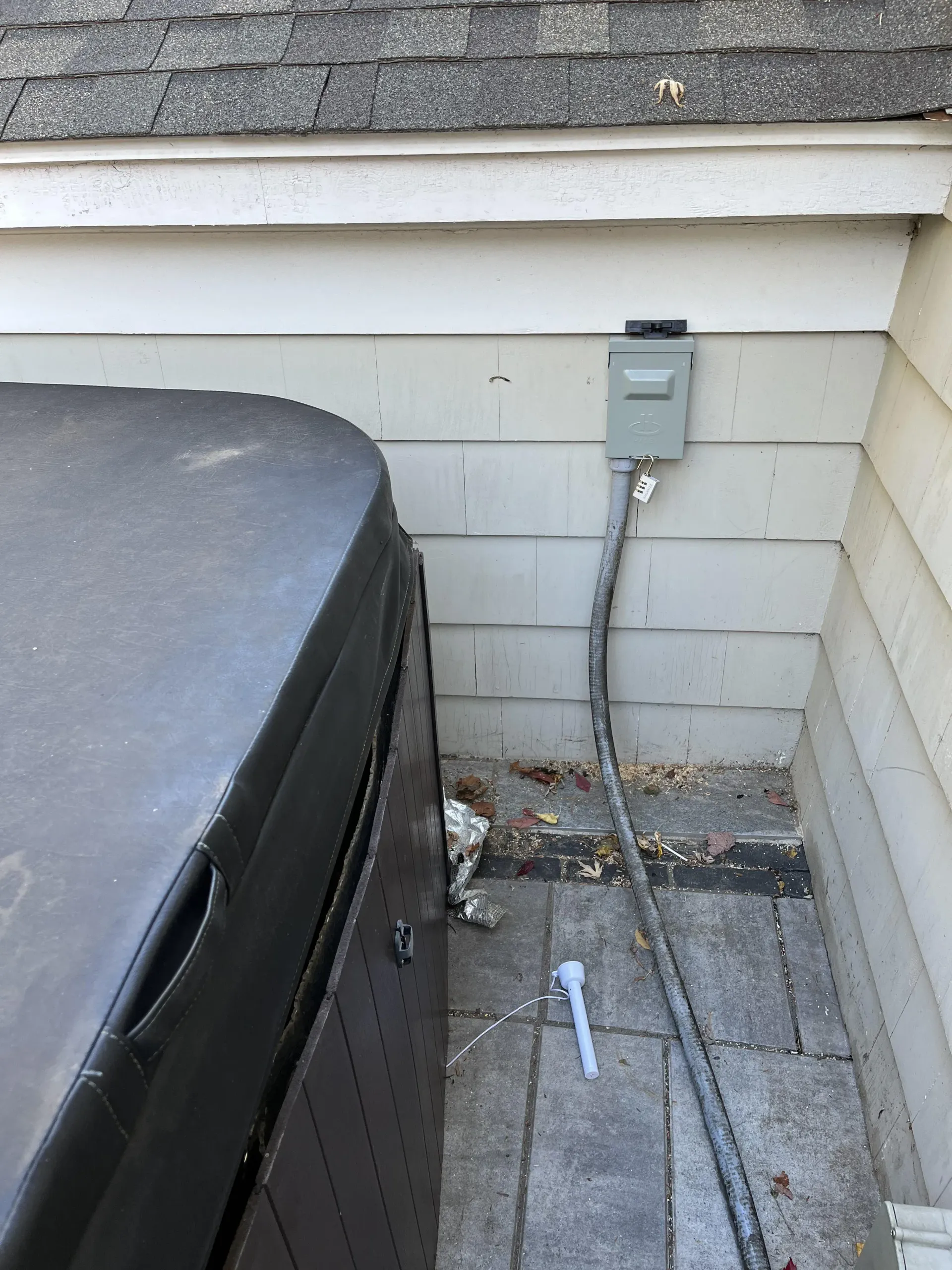Outdoor corner beside a house with an electrical box, utility meter, and trash bin on a concrete patio.