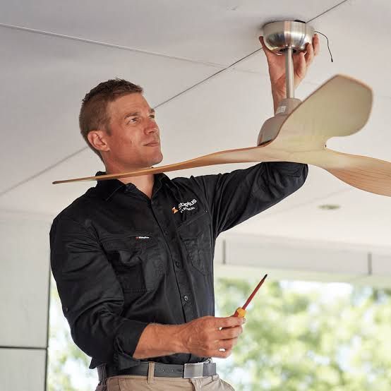 Electrician installing a ceiling fan, holding a screwdriver and looking up at the fixture.