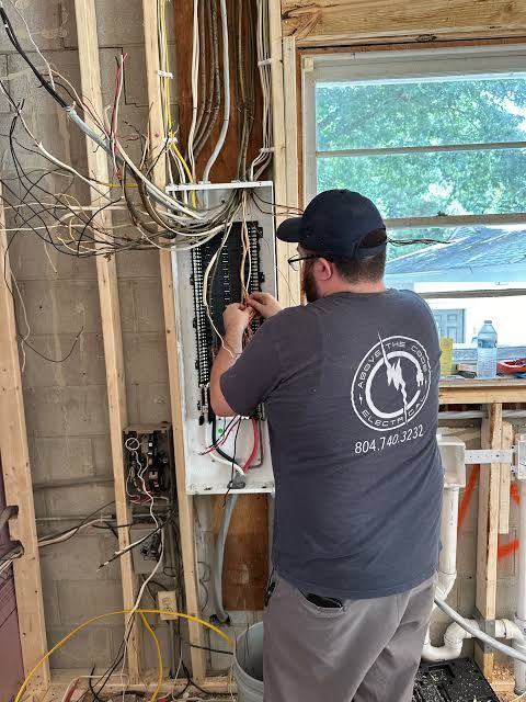 Worker wiring an open electrical panel in an unfinished room with exposed studs and cables.