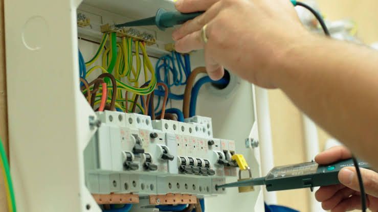 Electrician wiring a breaker panel with screwdrivers and colorful cables exposed inside an open electrical box