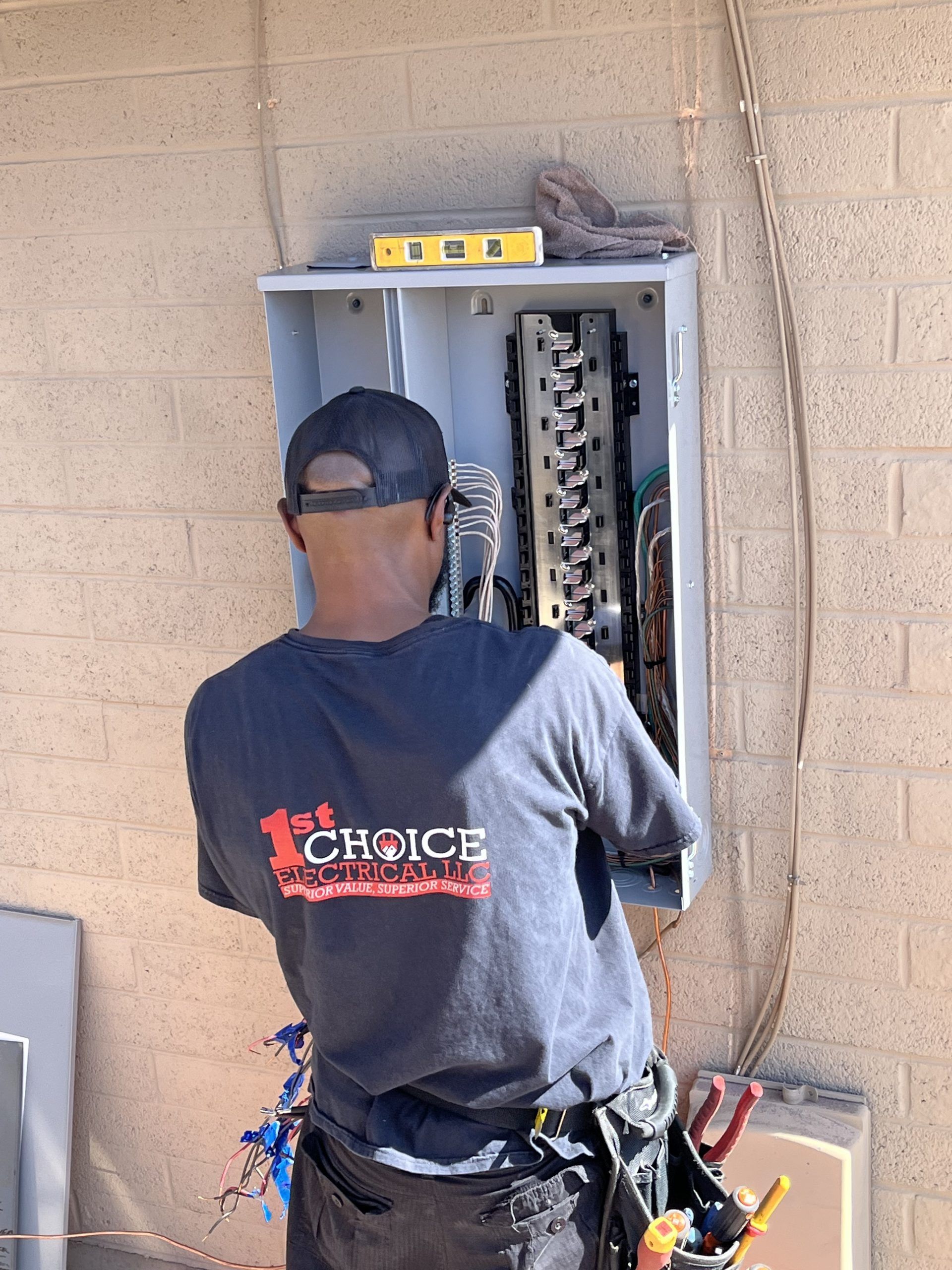Electrician working inside an open electrical panel on a beige cinderblock wall, wearing a tool belt.