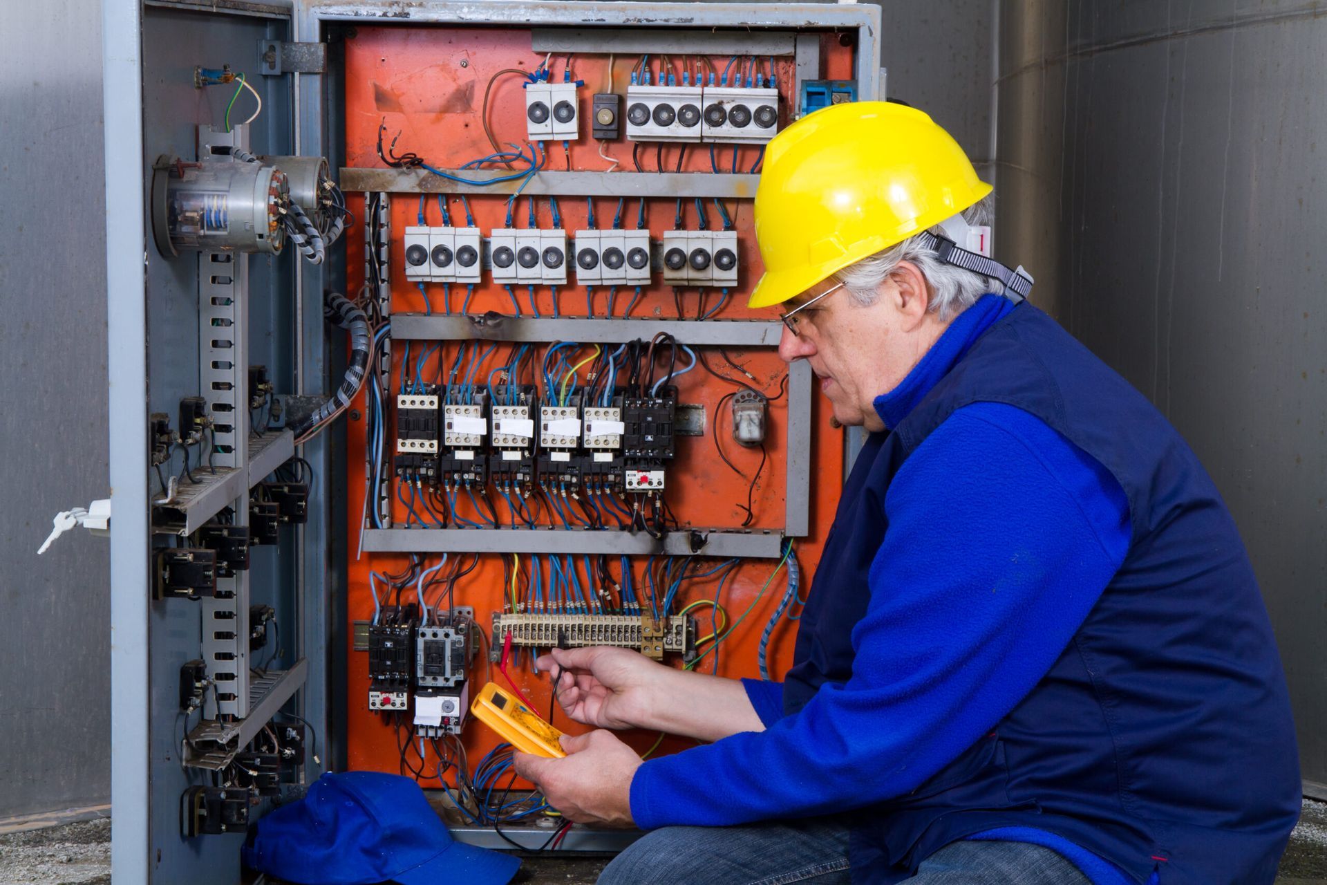 Electrician in yellow hard hat inspecting wiring inside an open electrical control panel
