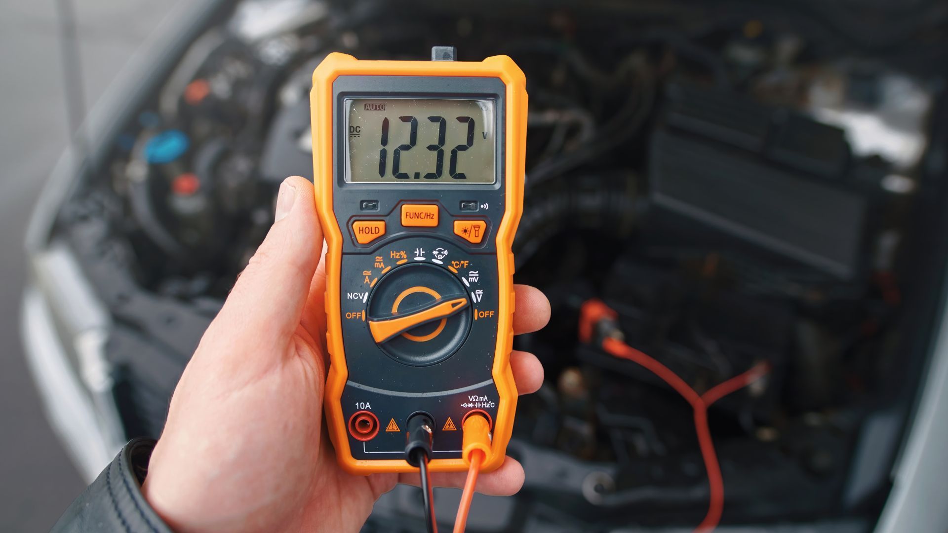 Hand holding a yellow multimeter showing 12.32 in front of an open car engine bay