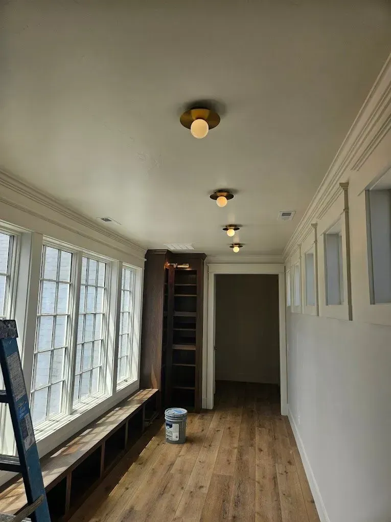 Long narrow room with windows on the left, wood floor, ladder, and a stairwell opening at the far end.