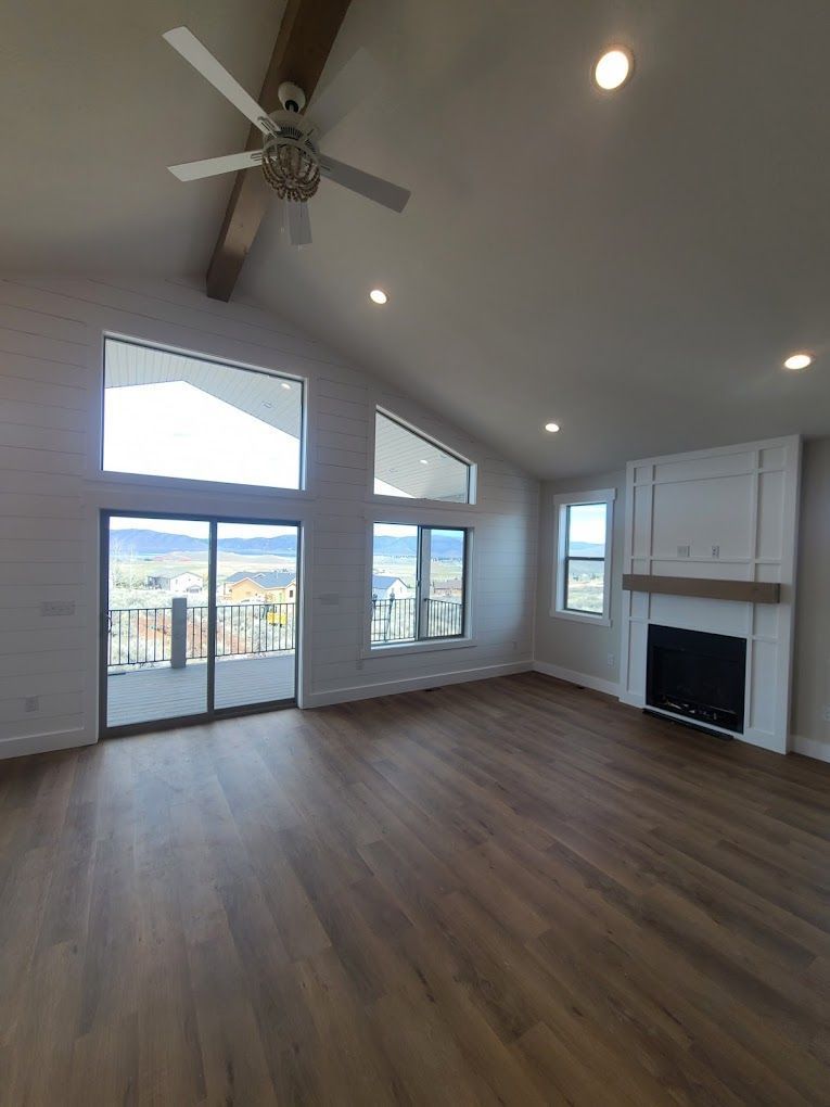 Bright empty living room with vaulted ceiling, ceiling fan, large windows, and fireplace.