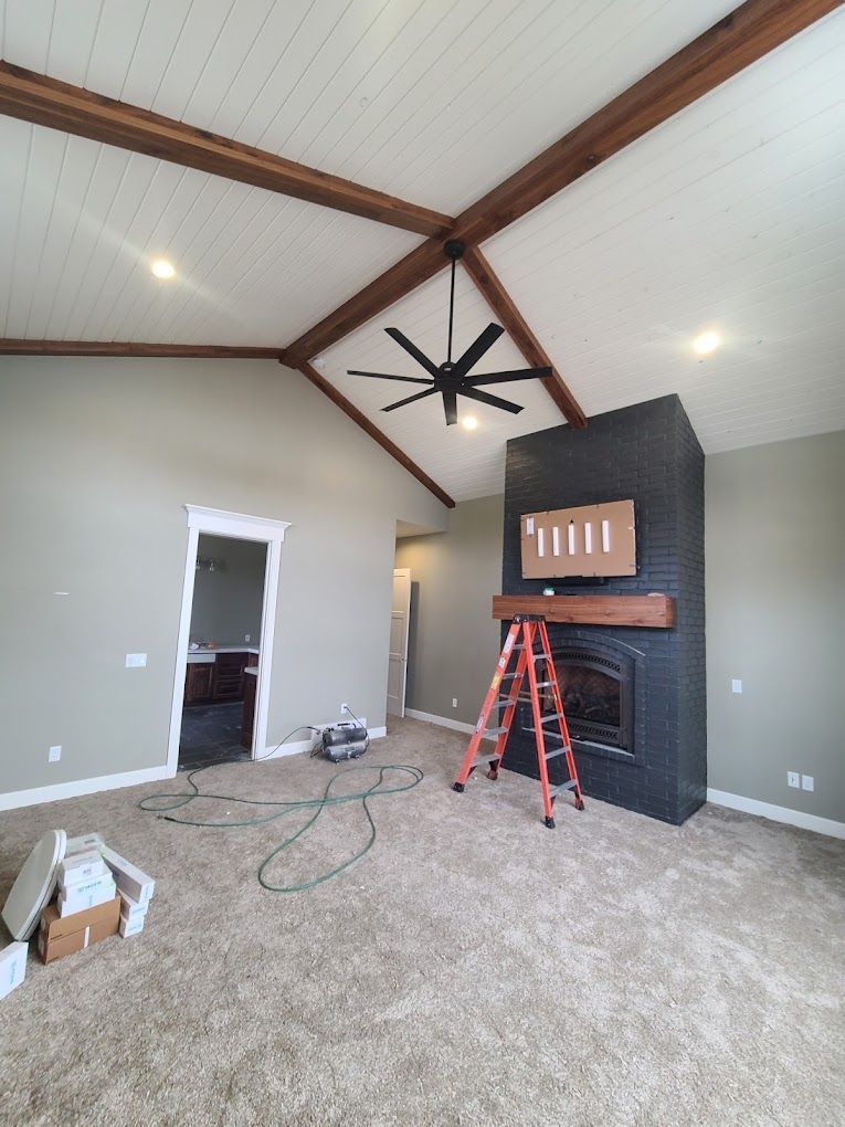 Living room under renovation with vaulted ceiling, black fan, fireplace, ladder, and tools on carpet