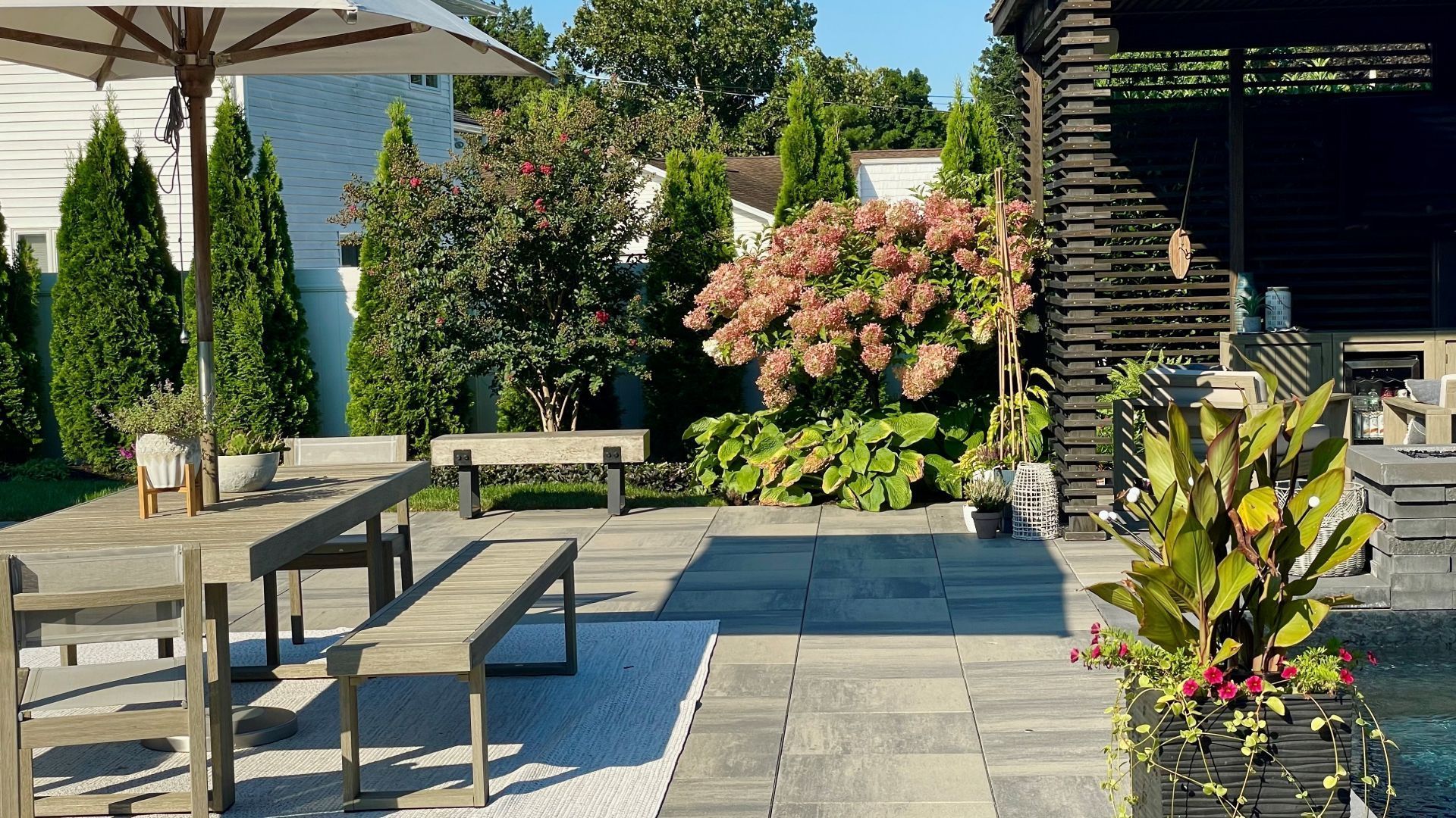 Sunny patio with stone pavers, outdoor seating, and lush potted flowers beside a garden pergola.