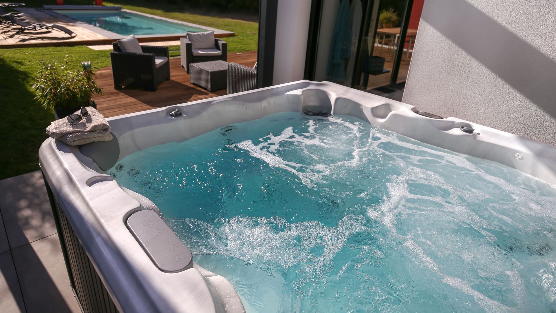 White hot tub with bubbling blue water on a sunny patio overlooking a pool and yard