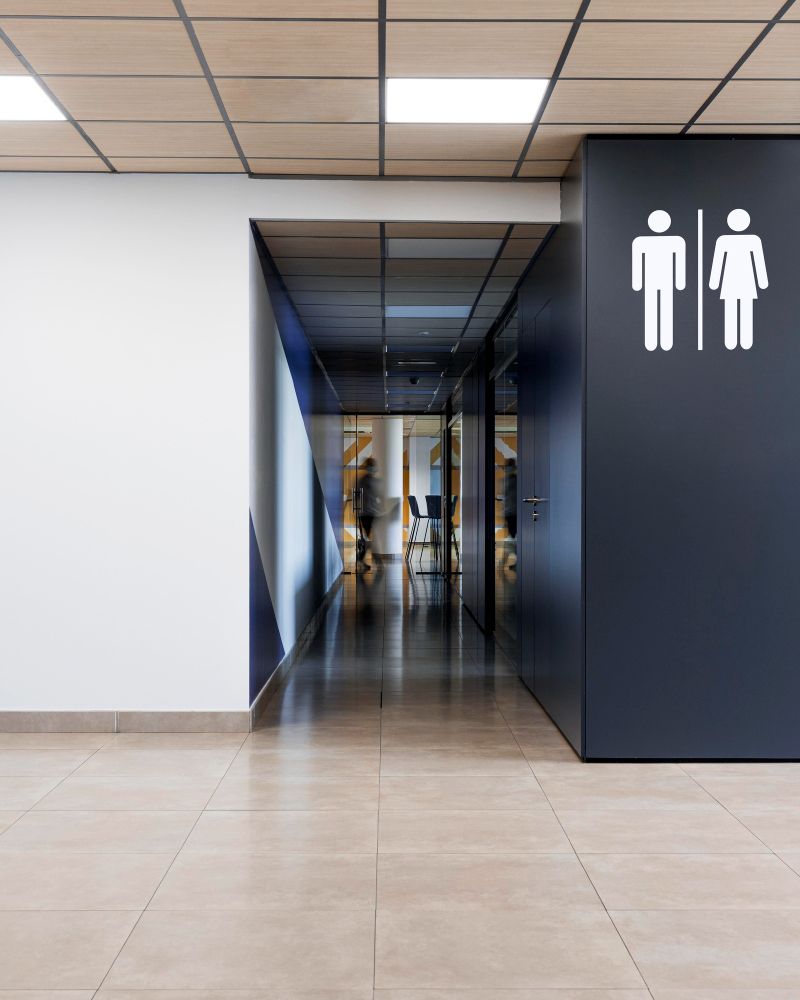Corredor moderno com paredes brancas e escuras, piso de azulejo e placas indicando o banheiro à direita.