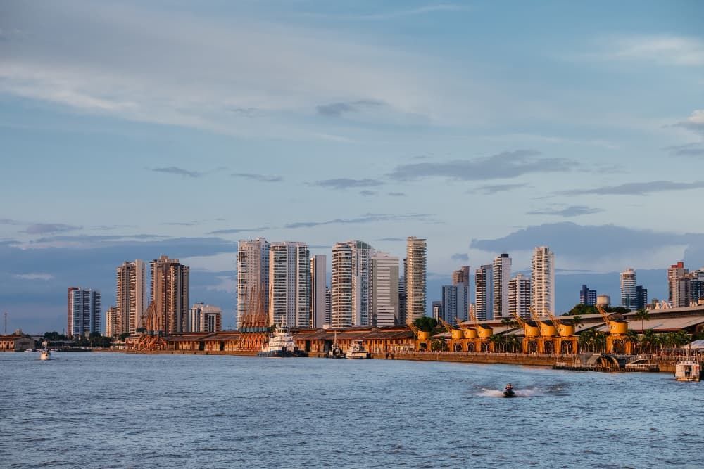 Vista da cidade ao entardecer sobre águas calmas, com barcos e uma orla repleta de prédios iluminados.