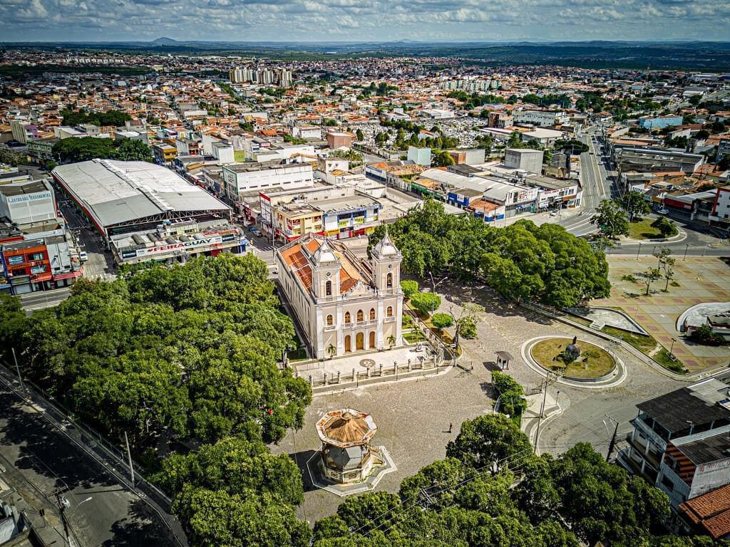 Vista aérea de uma praça da cidade com uma igreja histórica, árvores e edifícios circundantes sob um céu nublado.