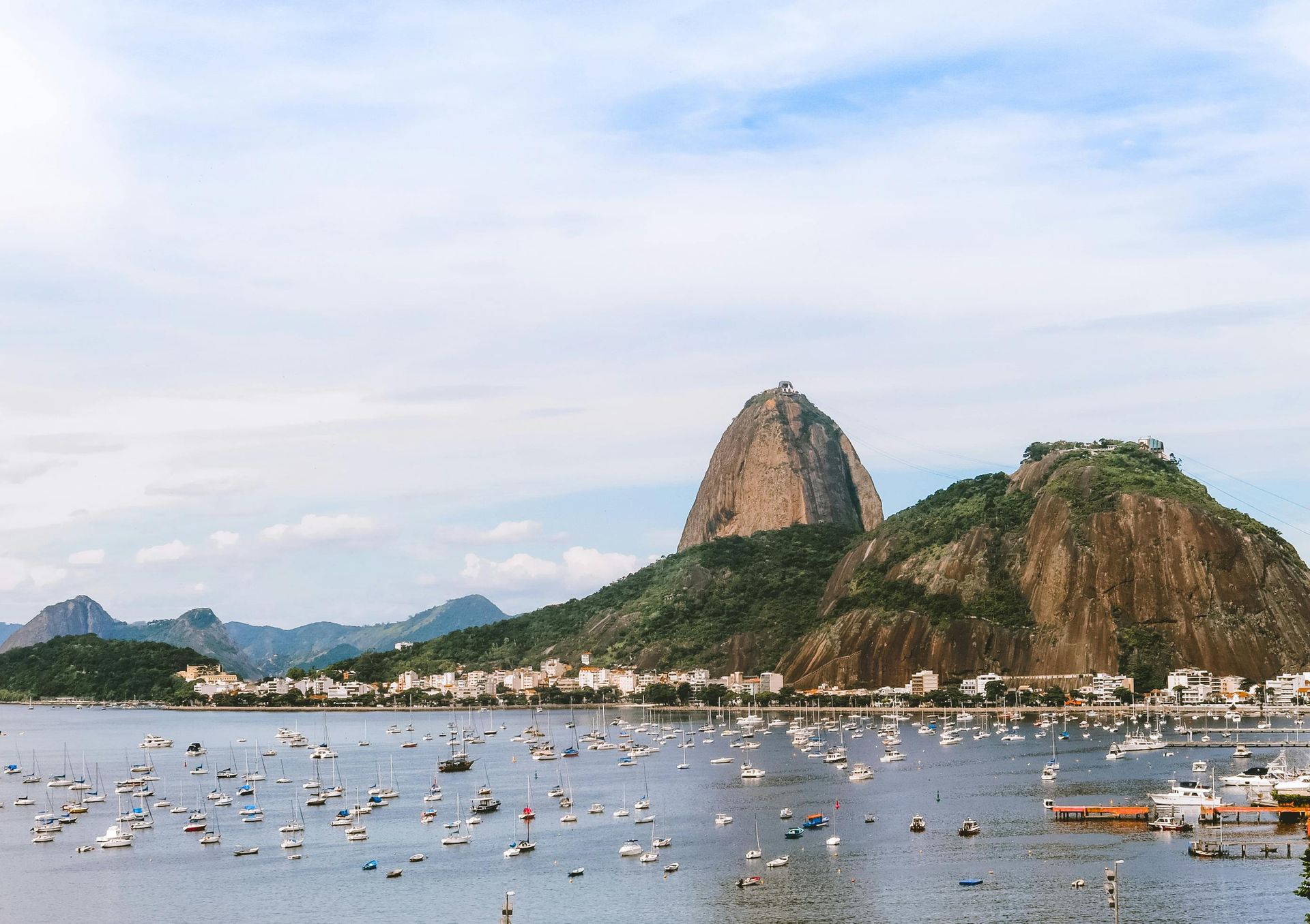 Baía costeira com barcos ancorados, montanhas verdejantes e um grande pico rochoso sob um céu nublado.