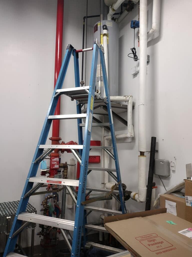 A blue ladder is sitting in a room next to pipes.