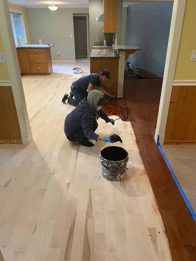 Two men are painting a wooden floor in a kitchen.