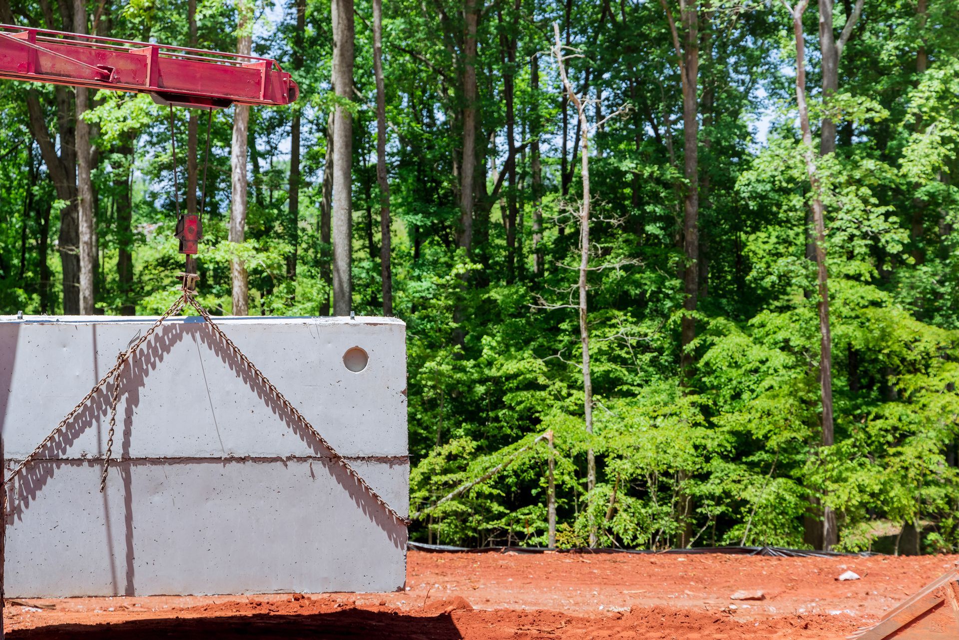 A concrete septic tank is being lowered into an excavated hole by a yellow excavator at a construction site.
