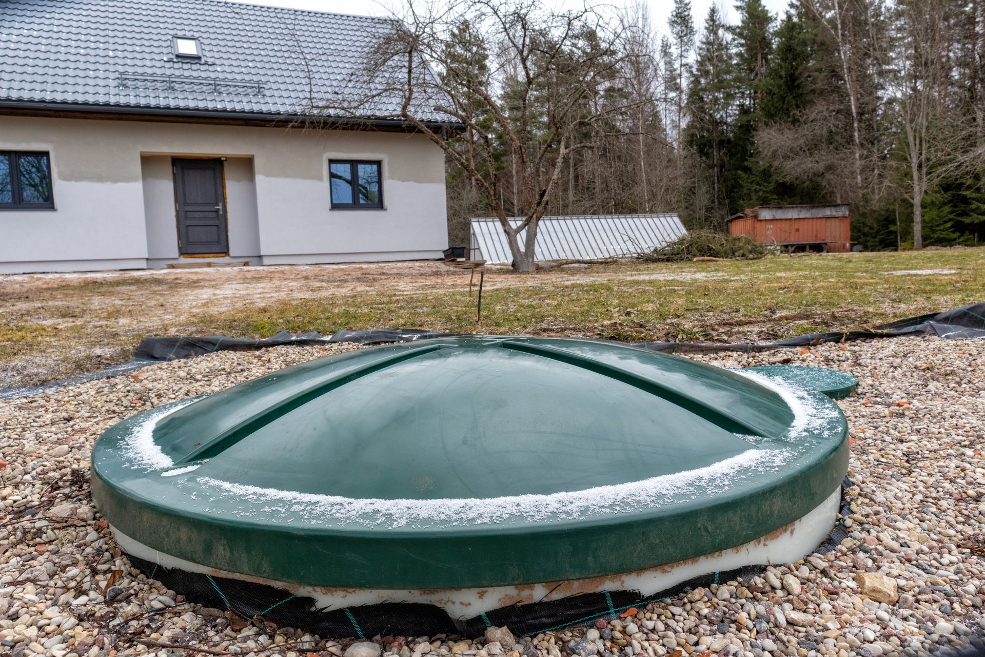 A green, dome-shaped septic tank cover in a gravel yard with a house and woods in the background.
