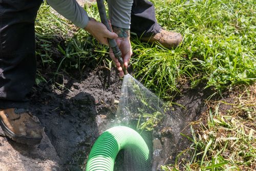 A worker in work boots sprays a water hose into a muddy trench near a large, green corrugated suction hose.