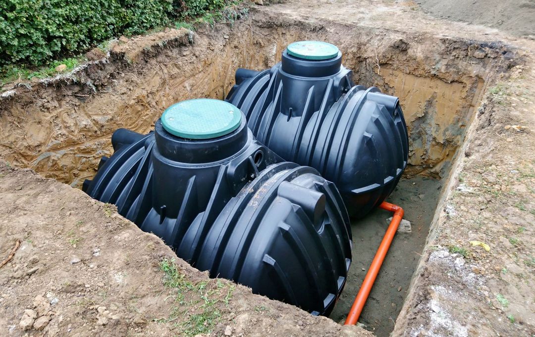 Two black plastic septic tanks installed in a dirt trench, connected by an orange pipe, with green access covers on top.