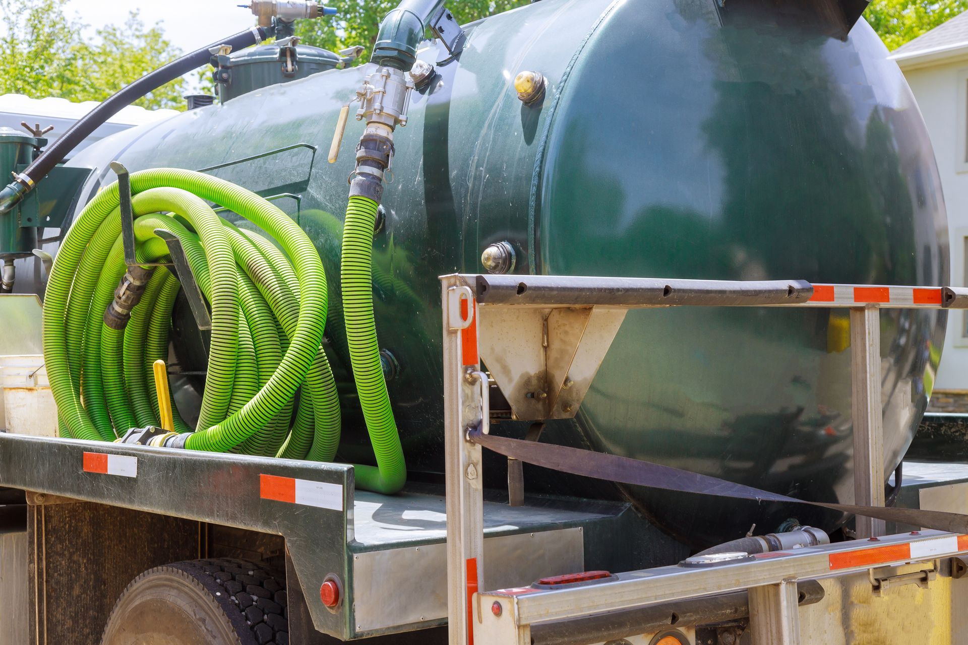 A large green tanker truck parked outdoors, featuring a coiled, bright green suction hose and red-and-white safety tape.