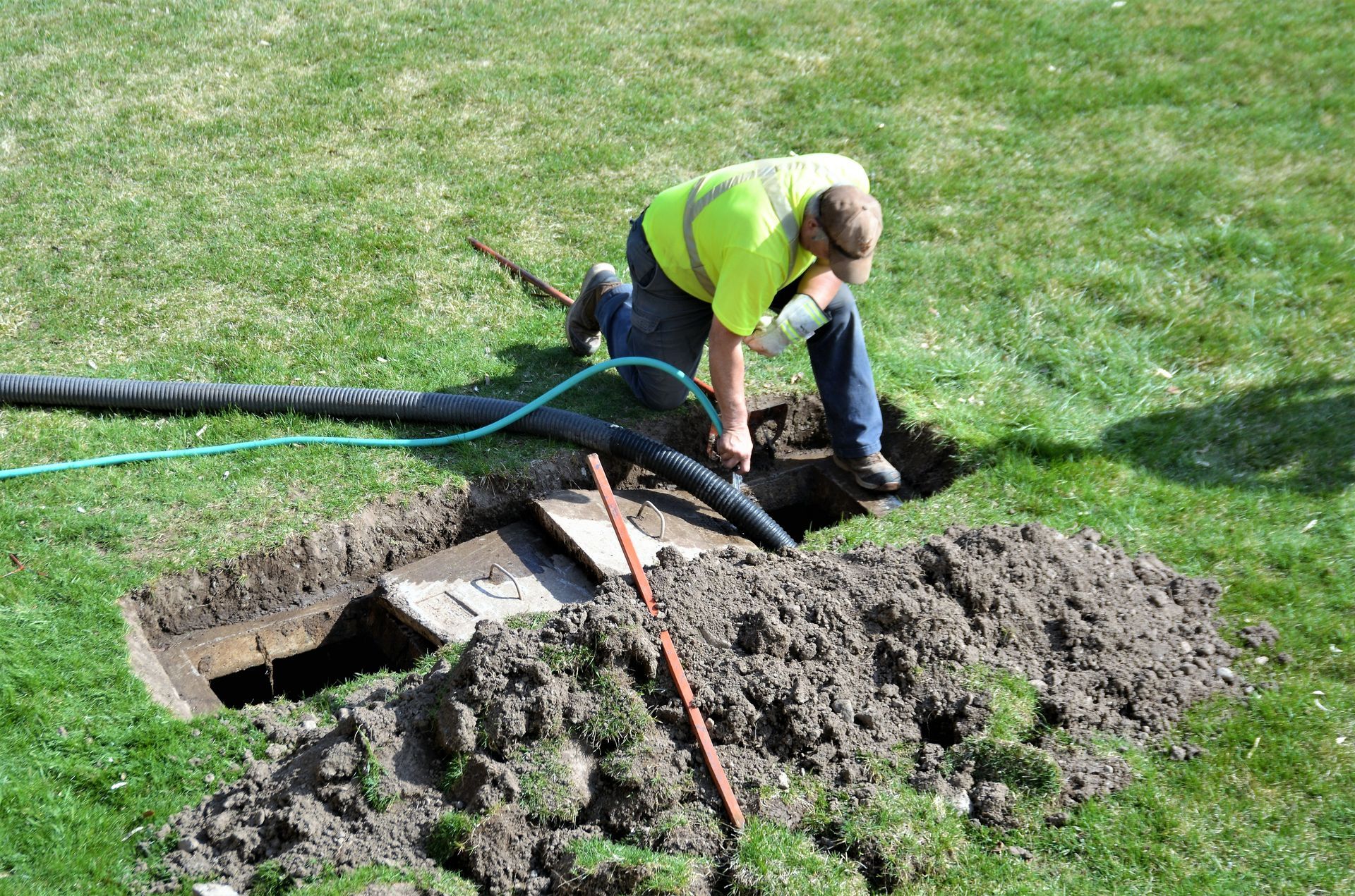 A worker in a high-visibility yellow shirt kneels in grass, using a black hose to excavate a narrow, rectangular trench.