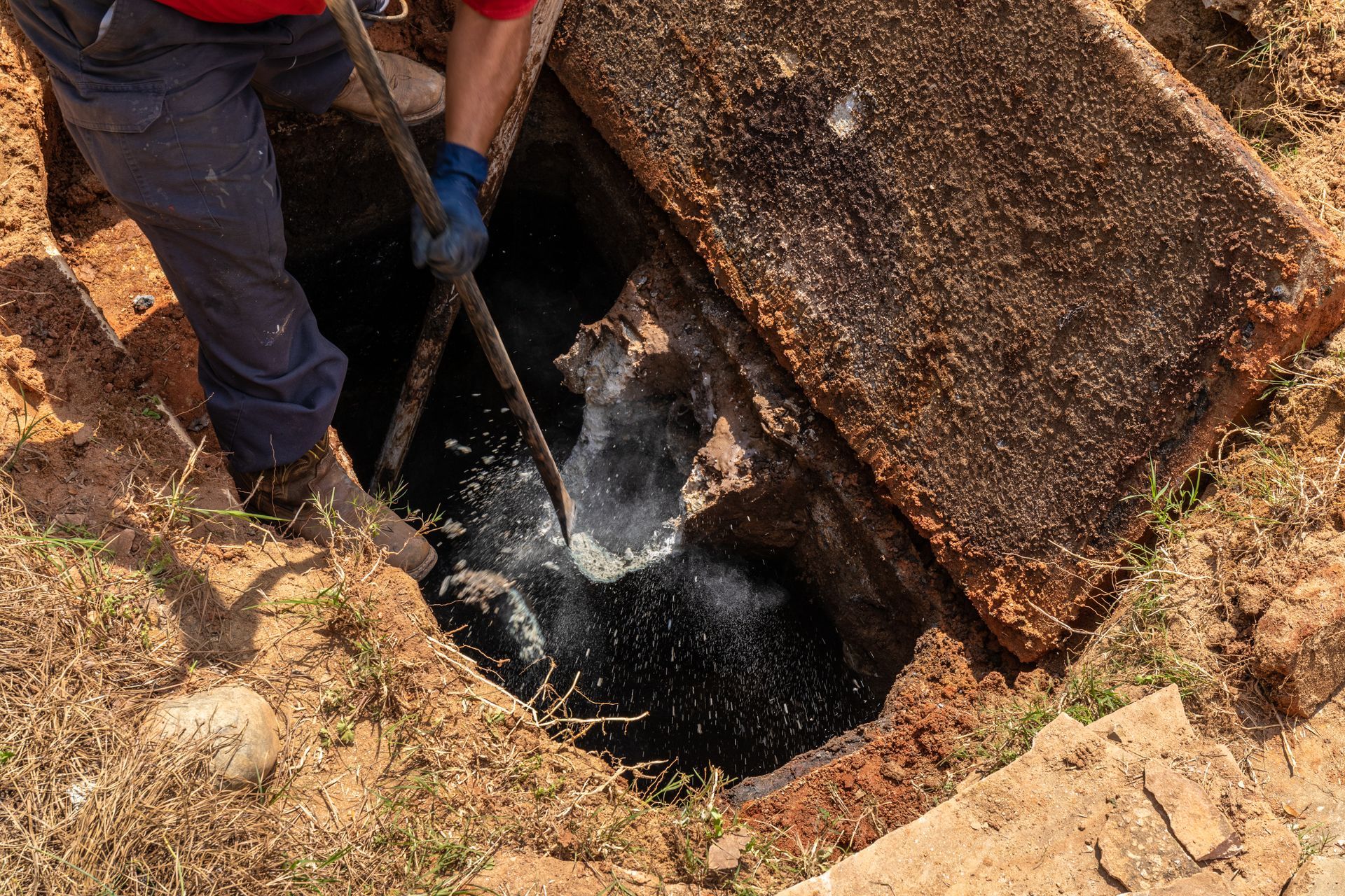 A person in uniform uses a long tool to clear a clogged, dark-colored drainage pipe in a dirt-lined trench.