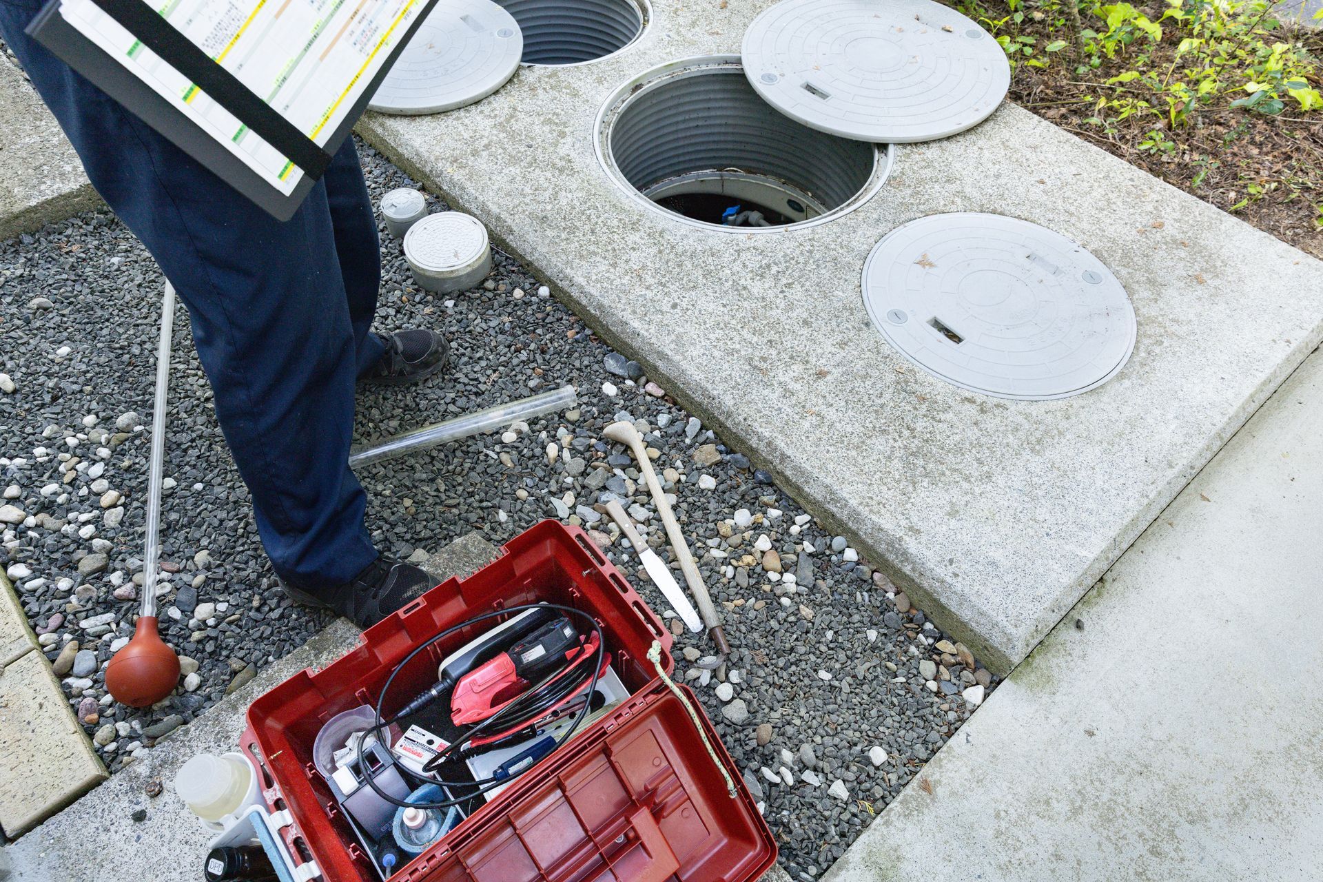 A technician stands over an open underground storage tank, holding a clipboard near a red toolbox filled with equipment.