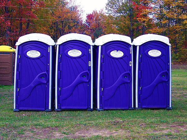 A row of identical blue portable toilets stands on a gravel lot under a clear, bright sky.