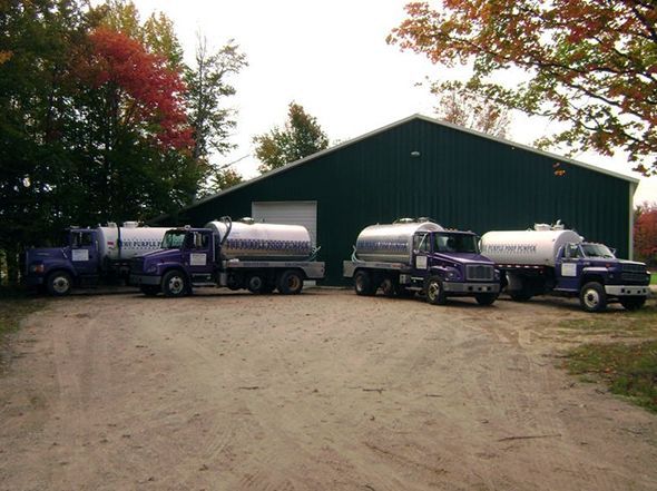 Four purple tanker trucks parked in a row on a gravel lot in front of a large, dark green storage building.