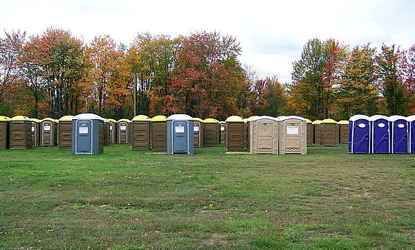 A row of dark green portable toilets with white roofs standing on a gravel surface under a clear blue sky.