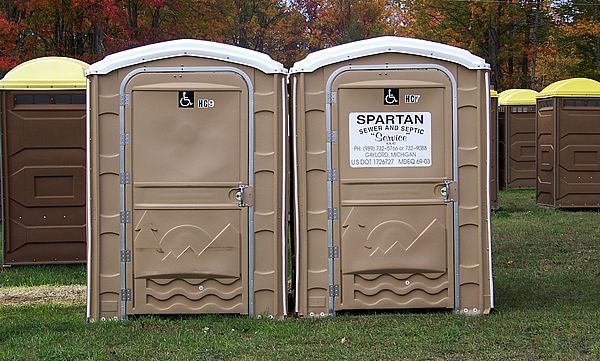 Two portable toilets, one light purple and one blue, stand side-by-side in a wooded area.