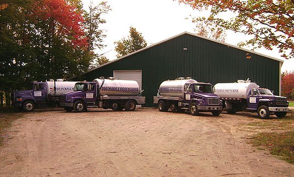 Four purple tanker trucks parked in a gravel lot in front of a green barn during autumn.