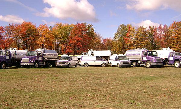 A line of purple utility trucks and white service vans parked in a field in front of a colorful autumn forest.