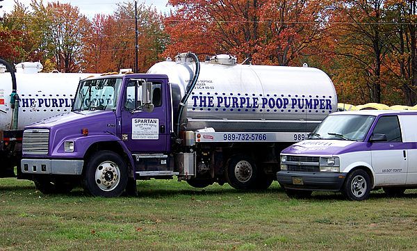 A worker wearing orange hi-vis trousers and gloves installs a circular black manhole cover set in fresh wet concrete.