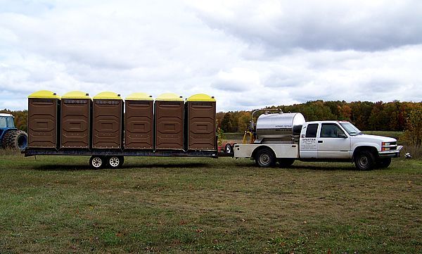 A white utility truck towing a long trailer loaded with six brown portable toilets in a grassy field under a cloudy sky.