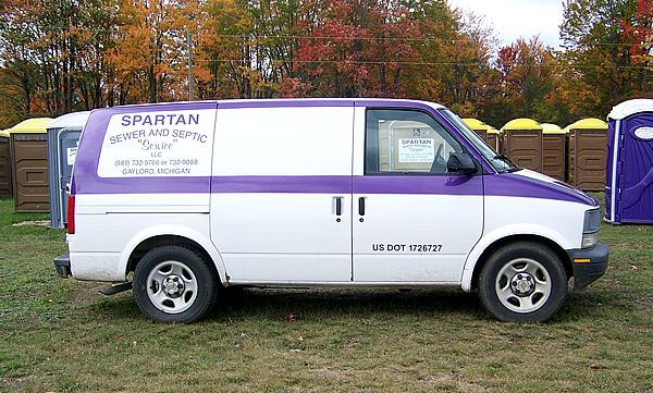 A white and purple Spartan portable toilet service van parked on grass in front of several portable toilets.