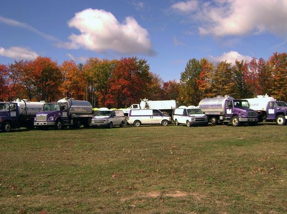 A line of white and purple service trucks parked on a grass field in front of a colorful autumn forest under a blue sky.